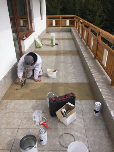 Handwerker verlegt Fliesen auf einer Terrasse, Werkzeug und Materialien liegen bereit.